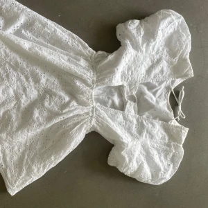 Vit kortklänning med puffärmar och broderade detaljer - Superfin vit kortklänning med puffiga ärmar och broderade blommönster. Klänningen har öppen rygg med knyt och en lätt volangig kjol. Perfekt för sommarens alla tillfällen!