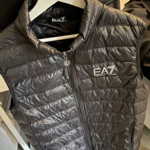 Svart dunväst från EA7 Emporio Armani - Snygg svart dunväst från EA7 Emporio Armani med dragkedja framtill och logga på bröstet. Västen har en quiltad design och hög krage, perfekt för lager-på-lager-stil.