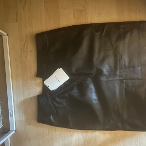 Svart minikjol i skinn från Gestuz - Säljer en svart minikjol i skinn från Gestuz. Kjolen har en stilren design med raka linjer och en ficka framtill. Perfekt för dig som vill ha en klassisk men edgy look.