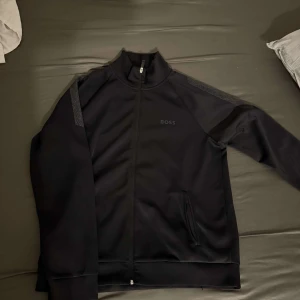 Svart track jacket från BOSS - Säljer en svart track jacket från BOSS med diskret logga på bröstet. Jackan har dragkedja framtill, ståkrage och två fickor med dragkedja. Snygga detaljer på axlarna och långärmad modell.