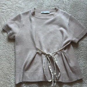 Beige stickad topp med knyt från Zara - Säljer en beige stickad topp från Zara med korta ärmar och rund halsringning. Toppen har ett fint knytband i midjan som ger en snygg detalj. Perfekt att styla till jeans eller kjol.