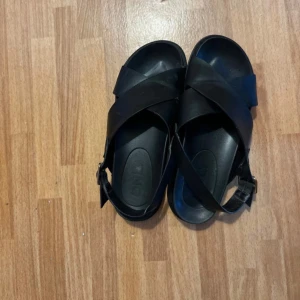 Svarta sandaler  - Säljer ett par svarta sandaler med breda korsade remmar och justerbart spänne vid hälen. Skorna har en platt sula och är tillverkade i syntetmaterial. Perfekta för varma dagar och enkel att matcha med olika outfits.