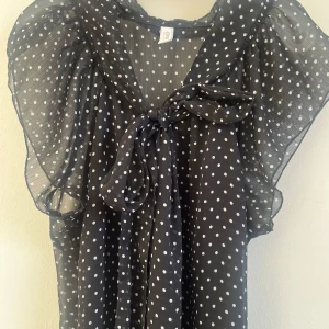 Svart prickig blus från ONLY - Säljer denna spuer snygga blusen från ONLY med vita prickar och volangärmar. Blusen har en fin rosettdetalj framtill och har även en litet svart tunnare tyg undertill. Perfekt för dig som gillar en klassisk och trendig stil.