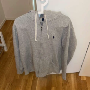 Grå hoodie från Polo Ralph Lauren - Säljer en klassisk grå hoodie från Polo Ralph Lauren med dragkedja och vit snörning i huvan. Tröjan har fickor framtill och den ikoniska lilla loggan broderad på bröstet. Perfekt för en avslappnad stil. Priset kan diskuteras 💕