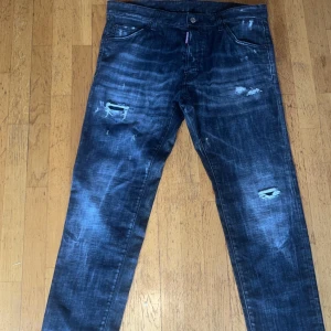 Mörkblå distressed jeans från Dsquared2 - Säljer ett par mörkgrå jeans från Dsquared2 med slitna detaljer och lätt tvättad look. Byxorna har klassisk femficksdesign, knappgylf och Dsquared2-logga på röd etikett vid fickan. Perfekta för en trendig streetstil.