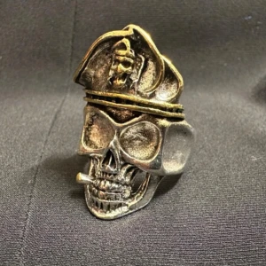 Ring i form av dödskalle med hatt - Cool ring i silverfärgad metall formad som en dödskalle med cigarett och detaljerad hatt med guldfärgade inslag. Perfekt statement piece för dig som gillar unika accessoarer med attityd.