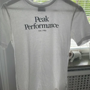 Vit t-shirt från Peak Performance - Vit t-shirt från Peak Performance med svart tryck på bröstet där det står 'Peak Performance Est. 1986'. Klassisk rund halsringning och korta ärmar. Perfekt basic-plagg med stilren design. I bra skick, köpt för 750kr! 🤍