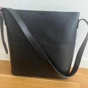 Svart axelväska i skinn - Snygg svart axelväska i äkta skinn med enkel och stilren design. Väskan har ett långt justerbart axelband och stängs med magnetknapp. Perfekt för att bära dina vardagsprylar med stil.