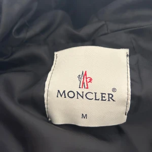 Svart pufferjacka från Moncler med huva - Säljer en svart pufferjacka från Moncler med huva och dragkedja. Jackan har broderad Moncler-logga på bröstet och stora vita Moncler-tryck längs huvan. Två sidofickor med dragkedja och quiltad design. Frakten står köparen för, eller hämtas från Borås.☺️