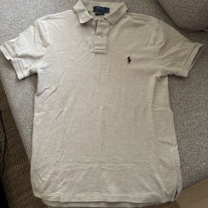 Beige pikétröja från Polo Ralph Lauren - Klassisk beige pikétröja från Polo Ralph Lauren med korta ärmar och krage med knappar. Ikoniska broderade loggan på bröstet. Perfekt för en stilren och avslappnad look. Knappt använd pga strl. 