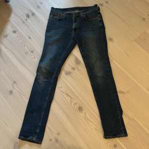 Mörkblå jeans från Nudie Jeans - Säljer ett par mörkblå jeans från Nudie Jeans med klassiska orange sömmar och raka ben. Jeansen har fem fickor och en snygg tvätt som ger en cool look. Perfekta för dig som gillar stilrena och tidlösa jeans.