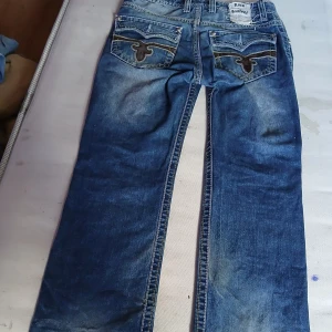 Rock revival Blå jeansbyxor med broderade bakfickor - Rock revival Säljer ett par klassiska blå jeansbyxor med snygga broderade detaljer på bakfickorna. Byxorna har normal passform och raka ben. Perfekta för en avslappnad stil och passar till det mesta i garderoben.