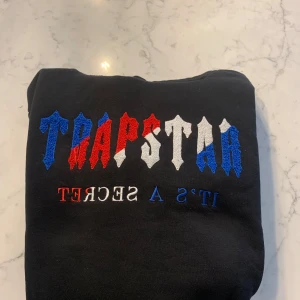 Svart hoodie från Trapstar - Säljer en svart hoodie från Trapstar med färgglad broderad text i blått, rött och vitt på framsidan. Hoodien har en klassisk design med dragsko i huvan och en stor magficka. Perfekt för en avslappnad stil. Priset kan såklart diskuteras!