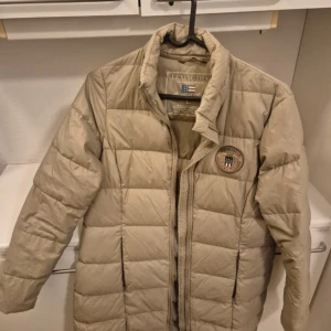 Beige dunjacka från Lexington - Säljer en snygg beige dunjacka från Lexington. Jackan har en quiltad design med ett broderat märke på bröstet. Den är långärmad och har en dragkedja framtill. Perfekt för kyligare dagar!