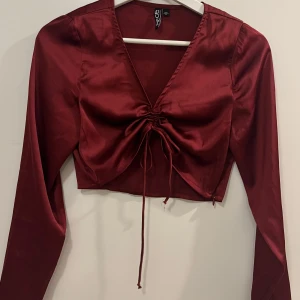 Vinröd satinblus från Pieces - Snygg vinröd blus i satin från Pieces med långa ärmar och en v-ringad design. Blusen har en knytning framtill som ger en fin detalj och en croppad passform. Samt dragkedja på sidan 