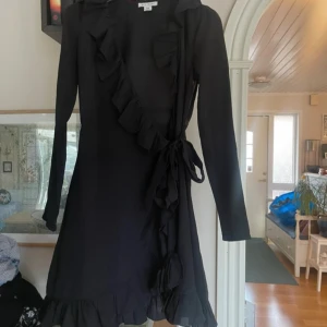 Svart omlottklänning från NLY Trend - Snygg svart omlottklänning från NLY Trend med volangdetaljer och knytning i midjan. Klänningen har långa ärmar och en elegant design som passar perfekt för olika tillfällen.