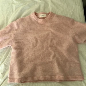 Rosa stickad tröja från Gina tricot  - Säljer en mjuk och stilren rosa stickad tröja från Gina tricot. Tröjan har en klassisk rund hals och korta ärmar, perfekt för lager på lager eller som den är. Den har en enkel design med ribbade kanter vid ärmslut och nederkant.