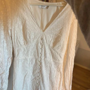 Vit klänning från Vila - Säljer en elegant vit klänning från Vila med v-ringning och långa ärmar. Klänningen har fina spetsdetaljer och knappar framtill. Perfekt för studenten!