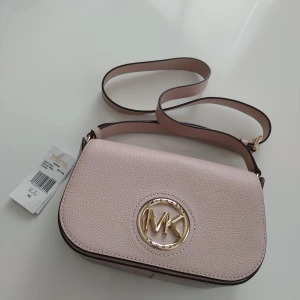 Rosa axelväska från Michael Kors - Säljer en stilren rosa axelväska från Michael Kors i skinn. Väskan har en elegant design med ett stort MK-emblem i guld på framsidan. Den justerbara axelremmen gör den bekväm att bära. Perfekt för att ge en touch av lyx till din outfit. Äkta läder och helt ny!