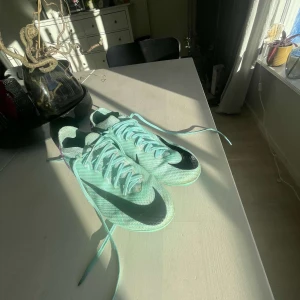 Nike Mercurial fotbollsskor i mintgrön och lila - Säljer ett par Nike Mercurial fotbollsskor i en fräsch mintgrön färg med lila detaljer. Skorna har en snygg svart Nike-logga och kommer med matchande skosnören. Perfekta för fotbollsplanen med sin lätta och smidiga design.