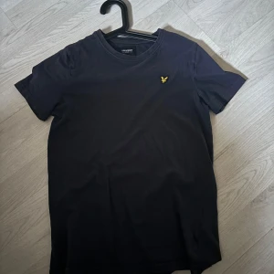 Svart t-shirt från Lyle & Scott Junior - Säljer en stilren svart t-shirt från Lyle & Scott Junior med korta ärmar. Den har en liten gul broderad logga på bröstet. Perfekt för en avslappnad look.