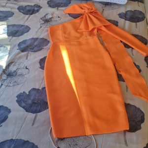 Orange axelbandslös klänning med rosett - Snygg orange klänning med en unik oneshoulder-design och en stor rosett vid axeln. Klänningen har en dragkedja baktill och är knälång, vilket ger en elegant look. Perfekt för festliga tillfällen!