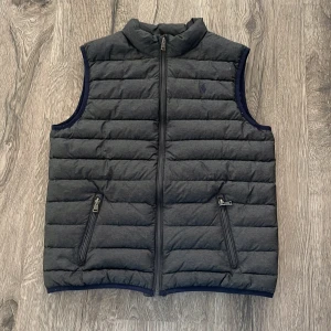 Ralph lauren väst - Köpt på zalando för 3500kr. Snygg grå quiltad dunväst från Ralph Lauren med dragkedja framtill och praktiska fickor med dragkedjor. Perfekt för lager-på-lager under kyliga dagar.