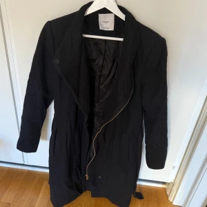 Svart vårkappa från Mango - Snygg svart vårkappa från Mango med dragkedja och knappar framtill. Den har ett bälte i midjan för en justerbar passform. Perfekt för kyligare dagar.