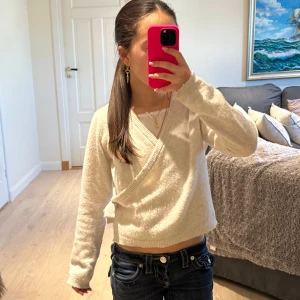 Vit omlottstickad tröja - Säljer en mysig vit omlottstickad tröja med långa ärmar. Perfekt för kyligare dagar och har en stilren design som passar till det mesta. Tröjan har en ribbad kant vid ärmslut och nederkant. Lite nopprig!