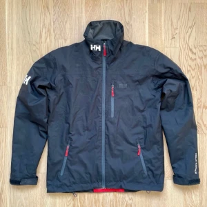 Svart vindjacka från Helly Hansen - Säljer en svart vindjacka från Helly Hansen med dragkedja och röda detaljer. Jackan har långa ärmar och flera praktiska fickor med dragkedjor. Perfekt för blåsiga dagar och har en stilren design med HH-logga på ärmen.