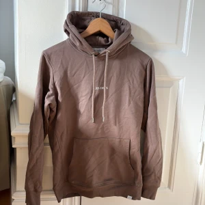 Brun hoodie från Les Deux - Säljer en stilren brun hoodie från Les Deux med justerbar huva och känguruficka. Perfekt för en avslappnad look. Den har en diskret logga på bröstet och är tillverkad i mjukt material.