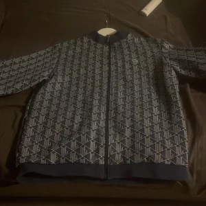 Mönstrad jacka från Lacoste - Säljer en snygg mönstrad jacka från Lacoste i blått och vitt. Jackan har en dragkedja framtill och långa ärmar. Perfekt för en stilren look kan även sänka från 600.
