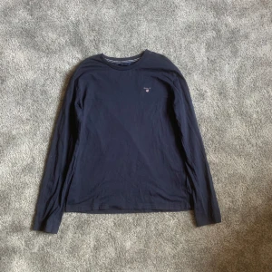 Mörkblå långärmad tröja från GANT - Säljer en stilren mörkblå långärmad tröja från GANT med broderad logga på bröstet. Perfekt för en avslappnad och snygg look. Passar bra till jeans eller chinos.