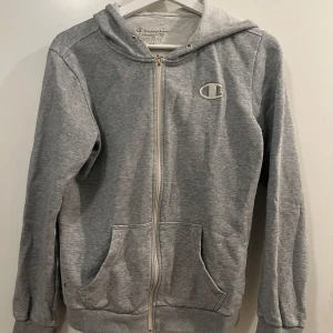 Grå hoodie från Champion - Säljer en klassisk grå hoodie från Champion med dragkedja och huva. Tröjan har långa ärmar och praktiska fickor framtill. 🩶