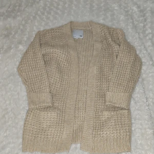 Beige stickad kofta från 157 - Mysig beige stickad kofta från 157. Perfekt för kyliga dagar med sin öppna design och långa ärmar. Koftan har ett fint stickat mönster och ribbade kanter som ger en stilren look.