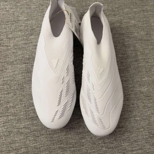 Fotbollsskor predator elite - Snygga vita fotbollsskor från Adidas med en stilren design. Skorna har en strömlinjeformad form utan snörning och är perfekta för spel på gräs. De har en lätt och flexibel konstruktion som ger bra grepp och kontroll på planen. Storlek 40 2/3