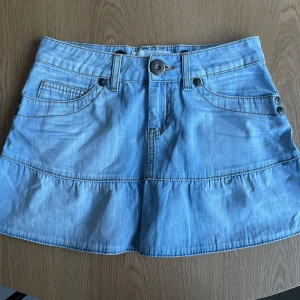 Ljusblå jeanskjol - Säljer en snygg ljusblå jeanskjol med dekorativa fickor och knappdetaljer. Kjolen har en kort längd och en volangdetalj nedtill. Perfekt för en avslappnad stil! Färgen ser ni bäst i sista bilden!