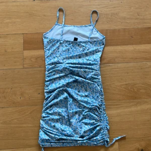 Blommig blå klänning från Shein - Säljer en söt blå klänning från Shein med blommigt mönster. Klänningen har smala axelband och en åtsittande passform med rynkade detaljer på sidorna. Perfekt för sommarens varma dagar! Längd justeras på sidorna! Använd väldigt lite, blir bara hängande! Ett hål i sidan lätt att sy ihop.