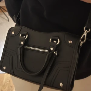 Svart väska - Snygg svart axelväska med silverdetaljer och dragkedja framtill. Väskan har både handtag och en justerbar axelrem för flexibel användning. Perfekt för att bära dina nödvändigheter med stil. Från HM men finns tyvärr inte kvar