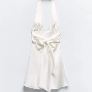 Vit student klänning  - Elegant vit halterneck klänning med en vacker rosett framtill. Klänningen har en djup v-ringning och är tillverkad i ett glansigt material som ger en lyxig känsla. Perfekt för sommarens festligheter.