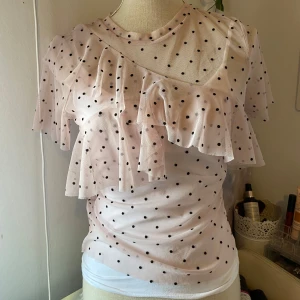 Rosa prickig blus med volang - Säljer en söt rosa blus med svarta prickar och volangdetaljer. Blusen har korta ärmar och är gjord i ett lätt och luftigt mesh material. Perfekt för en trendig look!
