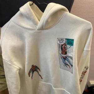 Vit hoodie med Spiderman-tryck - Säljer en cool vit hoodie med Spiderman-tryck. Den har en stor ficka framtill och tryck både fram och bak med serietidningsmotiv av Spiderman. Perfekt för fans av superhjältar! 🕷️