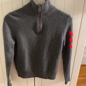 Grå tröja med dragkedja från Zadig & Voltaire - Snygg grå tröja från Zadig & Voltaire med dragkedja och ribbad krage. Den har röda detaljer på ärmen som ger en cool kontrast. Perfekt för kyligare dagar.