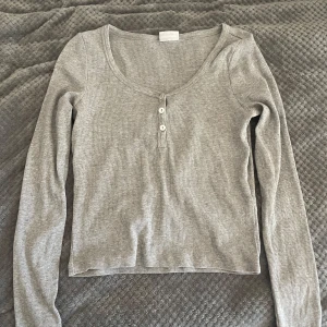 Grå ribbad tröja från Vera Moda - Säljer en stilren grå ribbad tröja från Vera Moda. Tröjan har en v-ringad design med tre knappar framtill och långa ärmar. Perfekt för en avslappnad look. Köpte för 300kr och använde bara det 2-3 gånger.