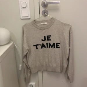Beige tröja med text från Gina Tricot - Säljer en beige tröja från Gina Tricot med texten 'JE T'AIME' i svart på framsidan. Tröjan har långa ärmar och en rund halsringning. Perfekt för en avslappnad stil.
