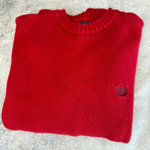 Röd stickad tröja från Chaps - Säljer en snygg röd stickad tröja från Chaps. Tröjan har en rund halsringning och ett diskret broderat emblem på bröstet. Perfekt för kyligare dagar och ger en stilren look.
