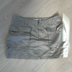 Beige kortkjol med fickor från 157 - Säljer en beige kortkjol från 157 med stora praktiska fickor framtill. Kjolen har en knapp och dragkedja i midjan samt hällor för bälte. Perfekt för en avslappnad stil.