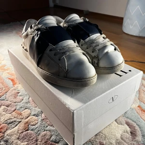 Valentino sneakers med svart detalj - Snygga Valentino sneakers i vitt med en bred mörkblå detalj över snörningen. Skorna har en klassisk design med rund tå och vita skosnören. Perfekta för en stilren look. skorna behöver limmas och har heal drag därav priset. pris är diskuterbart 