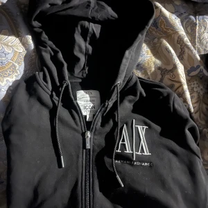 Svart hoodie från Armani Exchange - Säljer en stilren svart hoodie från Armani Exchange med dragkedja och huva. Den har en broderad logotyp på bröstet och praktiska fickor framtill. Perfekt för en casual look.