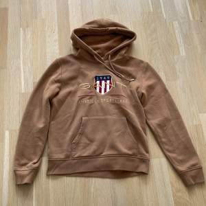 Säljer en snygg brun hoodie från GANT med ett stort tryck på framsidan. Hoodien har en känguruficka och justerbar huva. Perfekt för en avslappnad stil. Tillverkad i polyester för en mjuk och bekväm känsla.